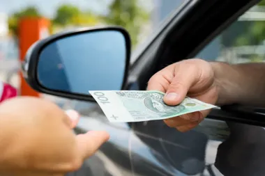 Kierowca wyciąga rękę z okna samochodu i wręcza banknot stuzłotowy drugiej osobie.